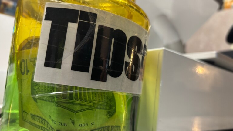 Closeup of a jar with a label that says “Tips” with cash inside.